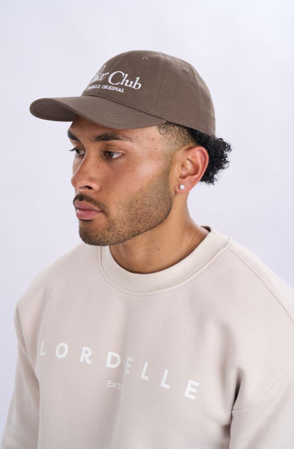 Man wearing a brown cap and beige sweatshirt with 'Lordelle' branding on a light grey background