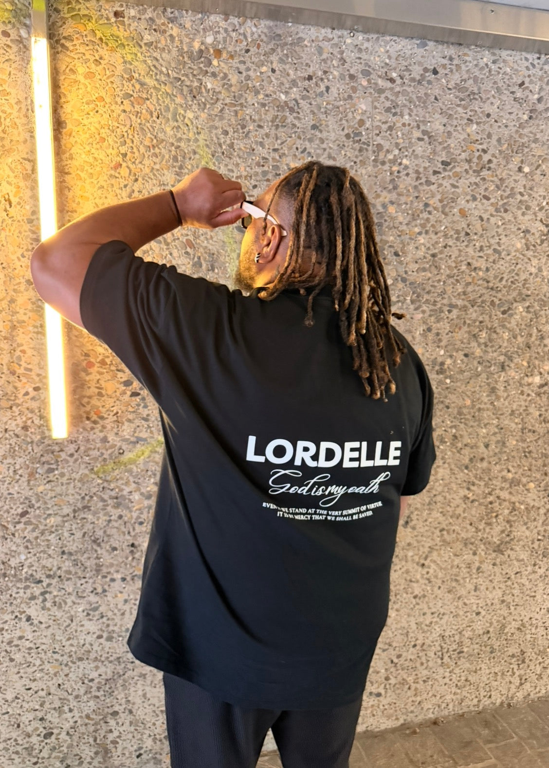 Person wearing a black t-shirt with 'LORDELLE' branding against a textured wall. God is my oath