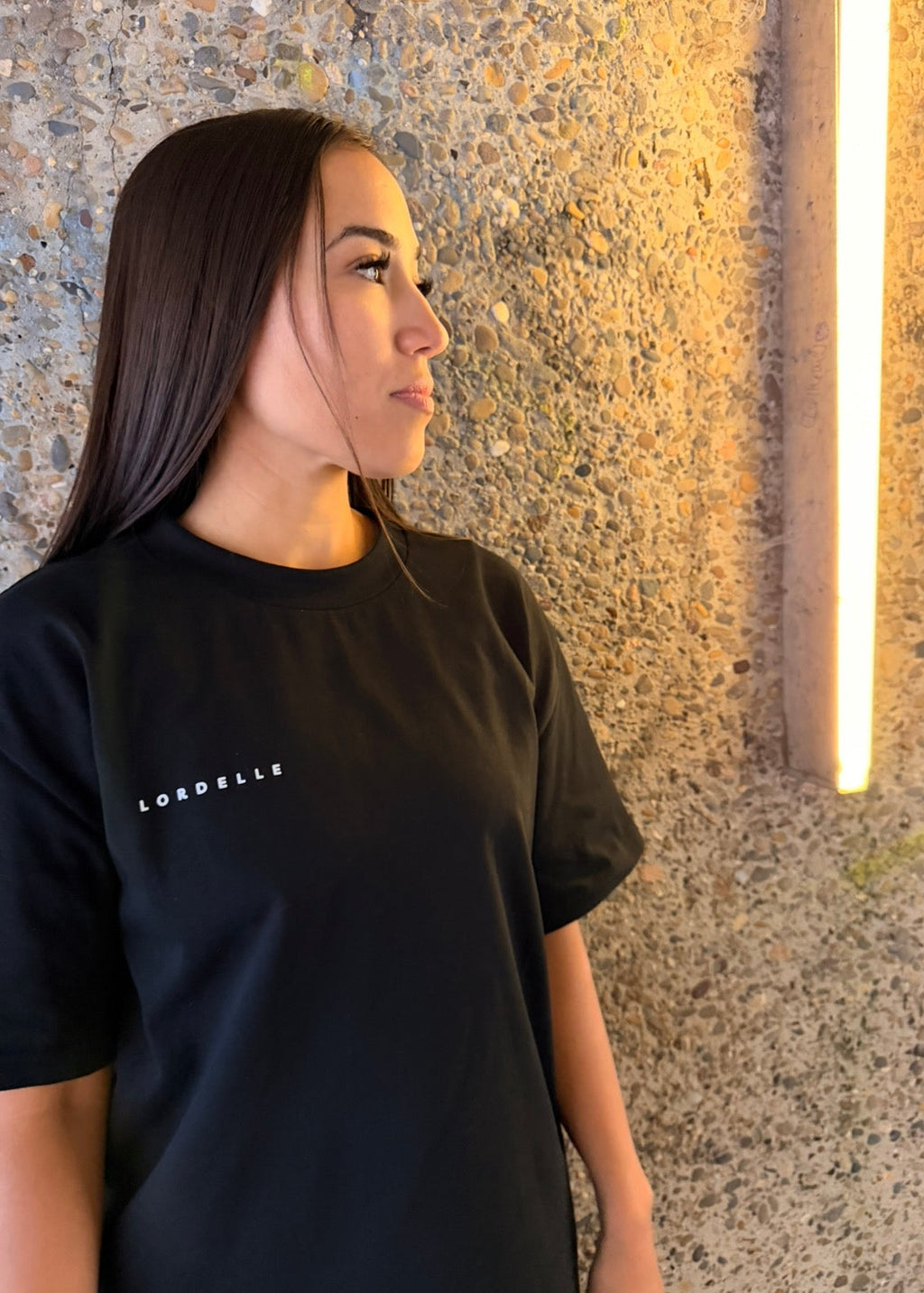 Person wearing a black t-shirt with 'Lordelle' logo against a textured wall.