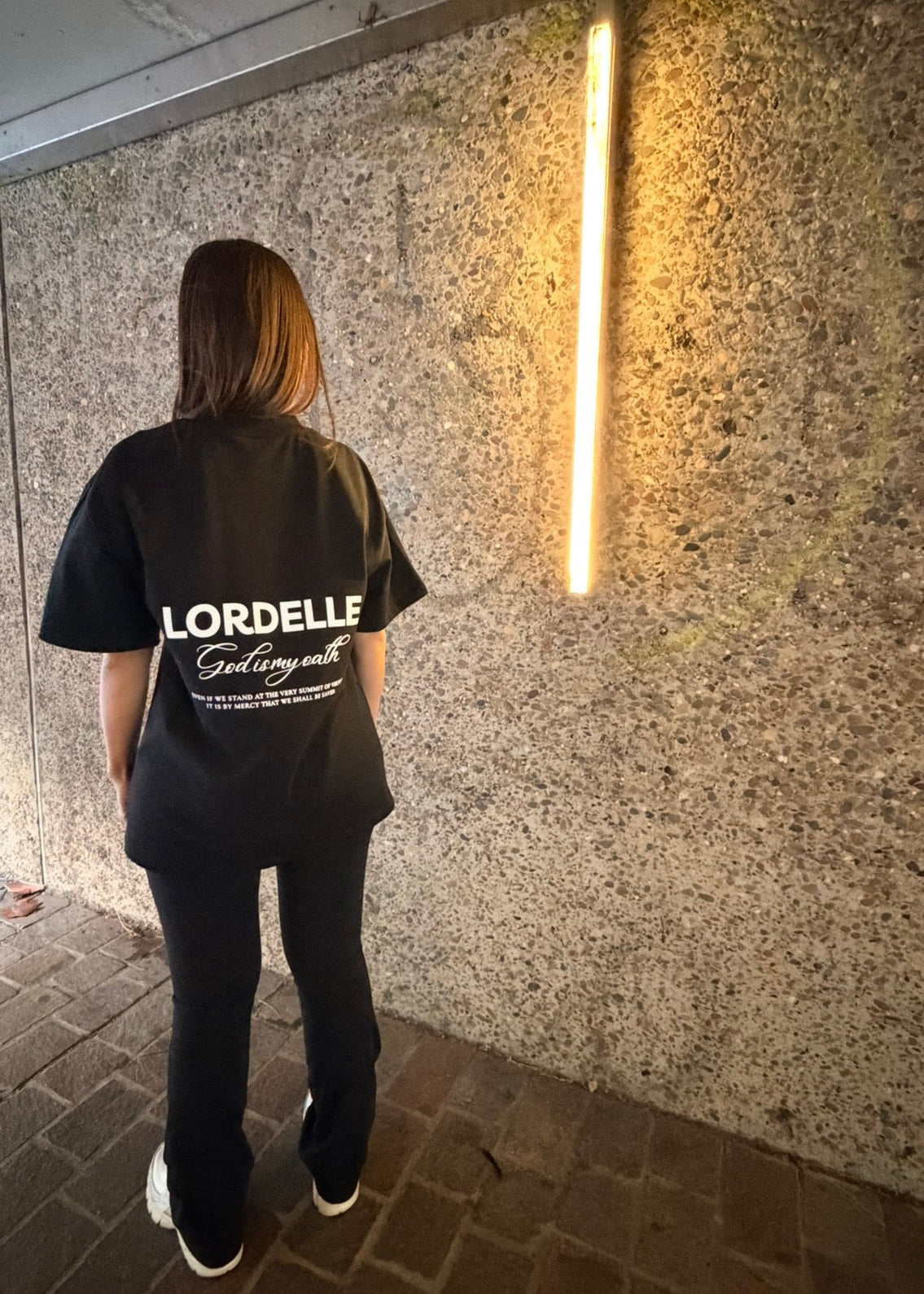 Person wearing a black t-shirt with 'LORDELLE' printed on the back, standing against a textured wall.
