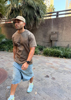 Man wearing a brown t-shirt and light blue shorts standing outdoors with plants and a wall in the background.