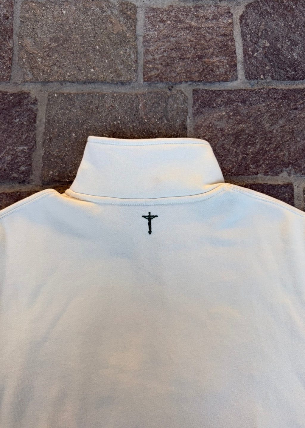 cream pullover with a black cross design on the back against a brick wall.