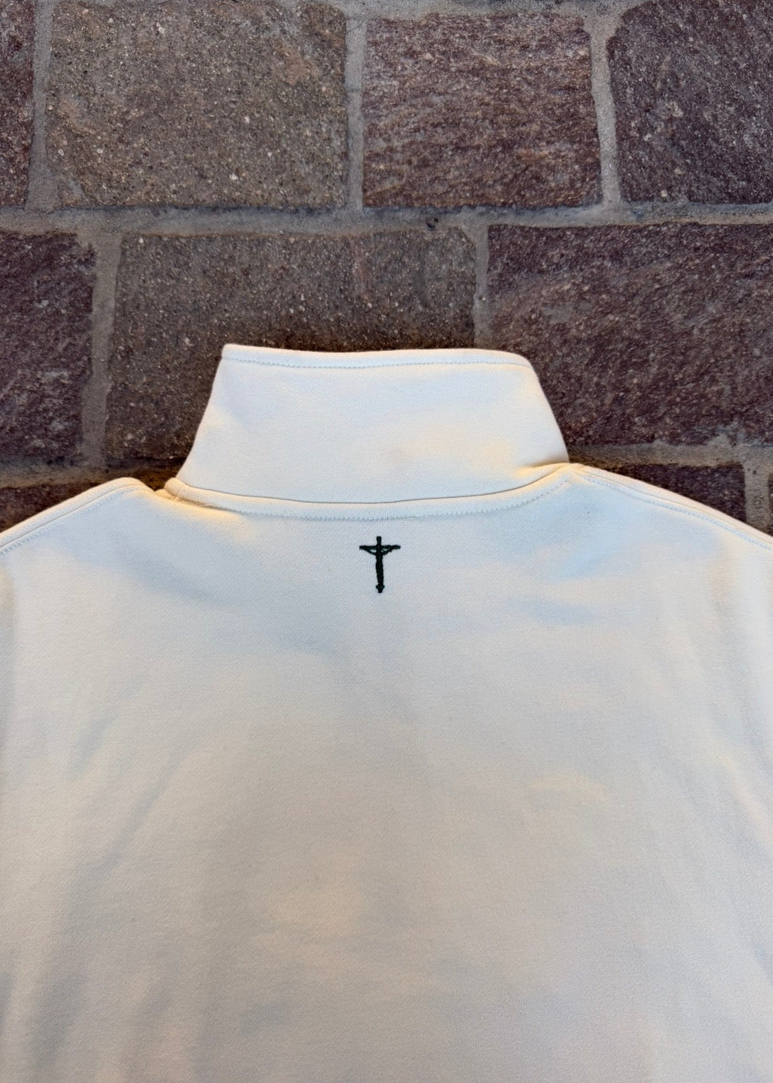 cream pullover with a black cross design on the back against a brick wall.