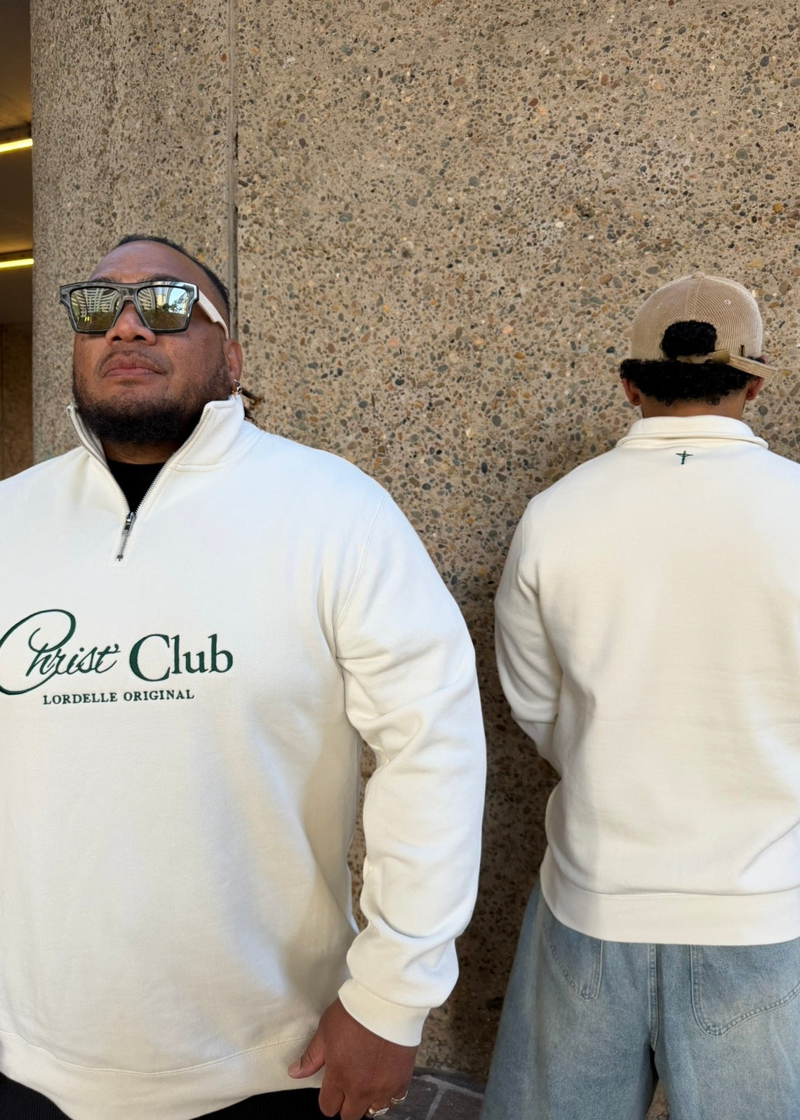 Two people wearing white sweatshirts with a logo, one facing forward and the other back, against a stone wall.
