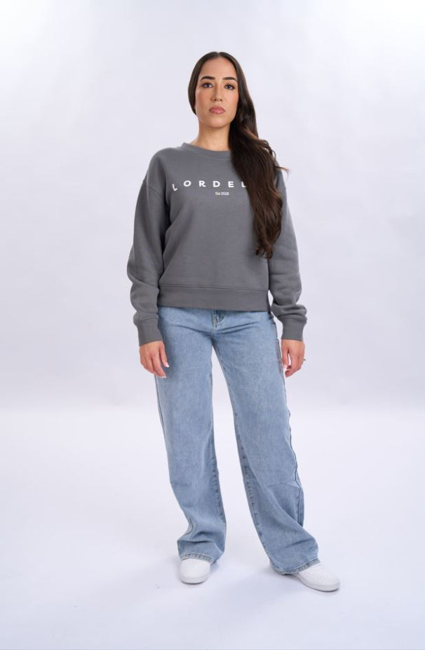Woman wearing a grey sweatshirt with 'LORDEN' branding and blue jeans on a white background